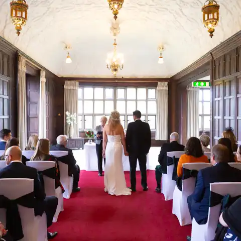 Wedding photo of the bride and groom and guests in the ceremony in the splendid Long Gallery at Fanhams Hall, Ware, Hertfordshire.  – photography by Glen Smith
