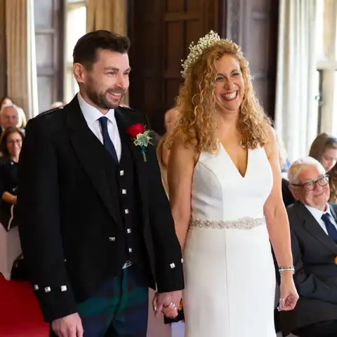 Wedding photo of the bride and groom holding hands in the Long Gallery in the ceremony and smiling at each other at Fanhams Hall, Ware, Hertfordshire.  – photography by Glen Smith