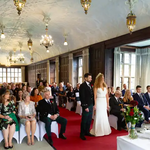 Wedding photo of the bride and groom and guests in the ceremony in the splendid Long Gallery at Fanhams Hall, Ware, Hertfordshire.  – photography by Glen Smith